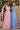 Two models wearing Ladivine formal gowns: one in a dusty rose pink one-shoulder dress with a flowing arm sash, and the other in a cornflower blue off-the-shoulder draped gown.