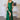 Model posing in Amoris emerald green formal dress (T817), featuring a ruched one-shoulder neckline, a fitted satin bodice, and a dramatic thigh-high slit with side cascade train.